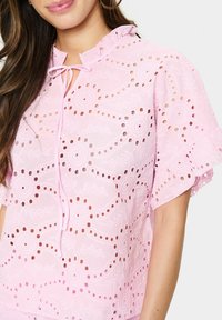 Top rosa in pizzo con colletto arricciato, caratterizzato da motivi floreali e design a taglio. Si lega al collo; maniche corte e svolazzanti.