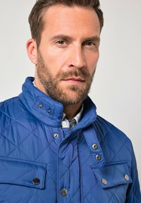 Blå quiltet jakke med høj krave, trykknapper og forlommer. Stoffet har en glat finish med diamantmønster.