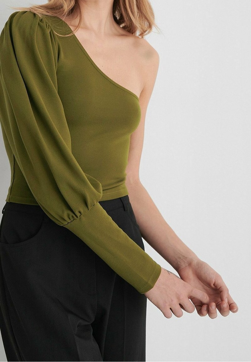 Femme portant un top vert olive à une manche avec une longue manche bouffante, associé à un pantalon noir taille haute sur un fond uni.