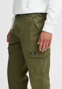 Olive green cargo pants with multiple pockets, features a side pocket with a label, made from durable cotton material, button closure.