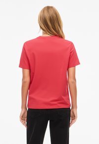 Pitkätukkainen nainen, jolla on yksinkertainen punainen lyhythihainen t-paita ja mustat housut, nähtynä takaa valkoista taustaa vasten.