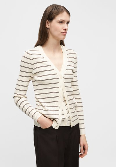 Γυναίκα που φοράει εκρού ζακέτα με λεπτές σκούρες οριζόντιες ρίγες, κουμπωμένη μερικώς, συνδυασμένη με μαύρο παντελόνι, σε πόζα με το ένα χέρι στην τσέπη.