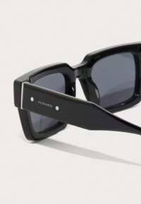 Melnas kvadr�āta formas saulesbrilles ar tumšiem stikliem, platām templēm ar sudraba akcentiem un zīmola "ALLSAINTS" nosaukumu sānos.