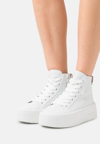 Bílé vysoké tenisky z hladké kůže s tlustou platformovou podrážkou, širokými tkaničkami a bočním zipem pro snadné nazouvání.