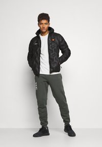 Schwarze Steppjacke mit gestepptem Design und angattached Kapuze, getragen über einem weißen T-Shirt. Olivgrüne Jogginghose mit weißen Akzenten und schwarzen Sneakers.
