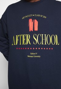 Felpa blu navy con stampe grafiche gialle e rosse. Presenta le scritte "AFTER SCHOOL", stelle e "Edition IV, Menance University".
