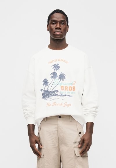 Άνδρας που φοράει λευκό φούτερ με εικόνα φοίνικα και κείμενο «Forever Paradise», μπεζ παντελόνι cargo, στέκεται μπροστά σε απλό φωτεινό φόντο.