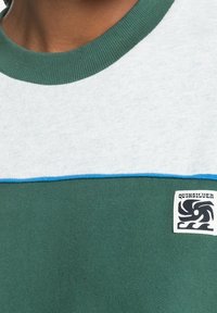 Lyhythihainen collegepaita, jossa on vihreä vartalo, harmaa yläpaneeli, sininen reuna ja musta logo-tagi. Valmistettu pehmeästä puuvillakankaasta.