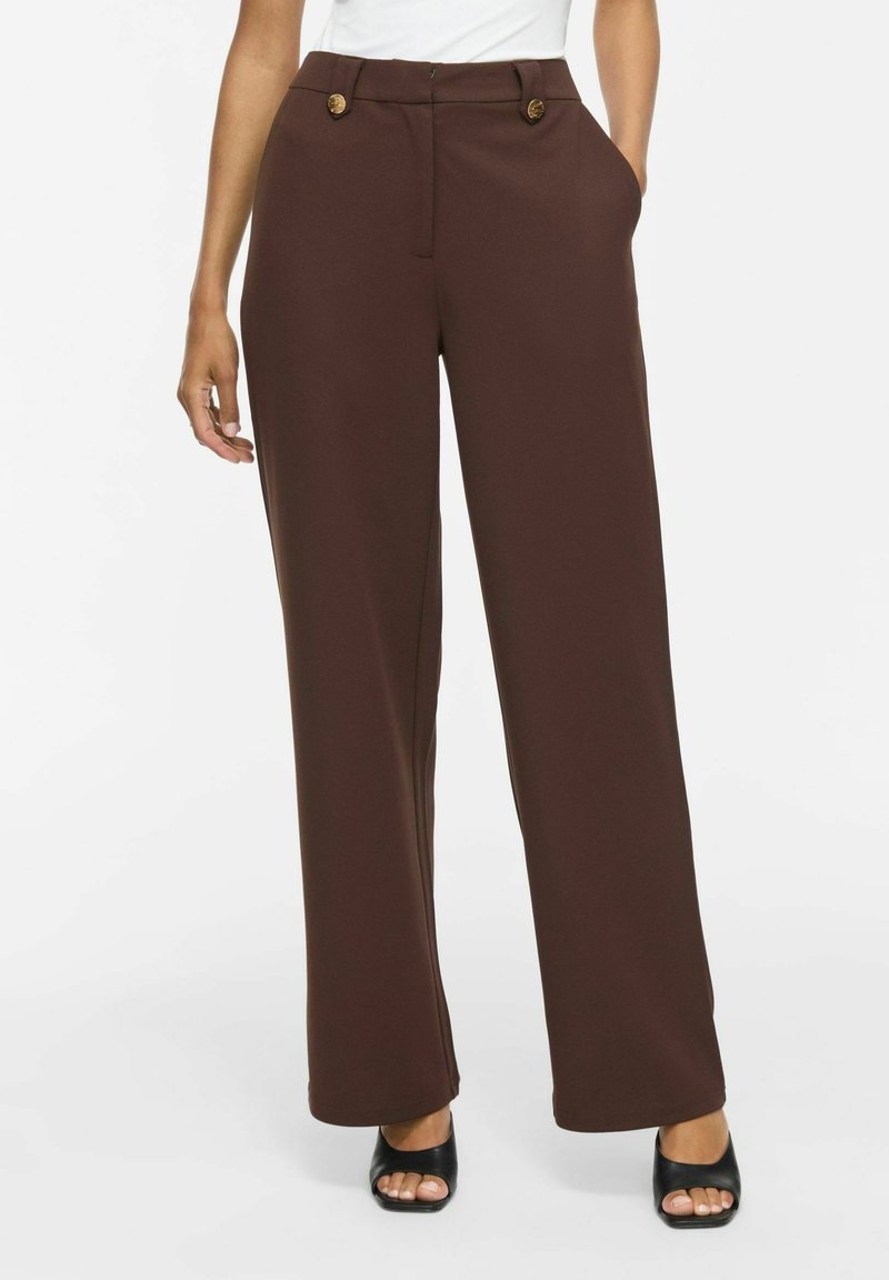 Pantalon marron à jambes larges avec des boutons à la taille, porté avec des sandales à talons noires à bout ouvert et un haut blanc.