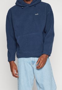 Felpa con cappuccio in pile blu navy con tasca a marsupio, cappuccio con coulisse e logo ricamato. Vestibilità comoda, superficie strutturata e maniche lunghe.