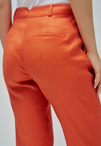 Primer plano de una persona que lleva pantalones naranjas de cintura alta con bolsillos traseros de ojal y una blusa blanca metida, con la mano descansando sobre el muslo.