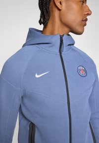 Felpa con cappuccio Nike di colore blu chiaro, con zip nera, tasche laterali e logo del Paris Saint-Germain sul petto, realizzata in tessuto testurizzato.