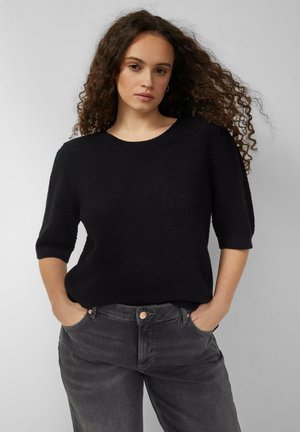 Junge Frau mit lockigem Haar, die einen schwarzen, texturierten Kurzarmpullover und dunkelgraue Jeans trägt, Hände in den Taschen.