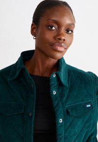 Veste en velours côtelé teal avec un devant boutonné, une fermeture à pression, une poche poitrine et un motif matelassé. Portée par-dessus un haut noir.