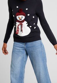 Persona che indossa un maglione nero con un pupazzo di neve sorridente con un cappello rosso e una sciarpa, abbinato a jeans larghi blu chiaro.
