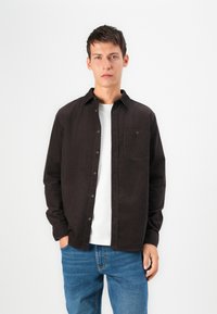 Camisa negra de botones hecha de tela texturizada, con un único bolsillo en el pecho, superpuesta sobre una camiseta blanca, combinada con jeans de mezclilla azul.