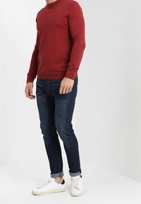 Czerwona koszula z długim rękawem, niebieskie jeansy o dopasowanym kroju oraz białe trampki. Koszula ma okrągły dekolt; jeansy są podwinięte na dole dla stylu.