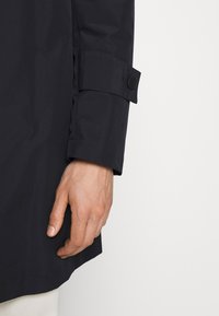 Manteau imperméable noir avec un poignet à boutons, présentant une texture lisse et un ourlet droit, conçu pour une coupe ajustée.
