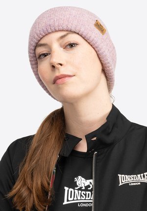 Stickad rosa mössa med ribbad textur och en liten läderetikett. Bärs tillsammans med en svart jacka med dragkedja och logotyp på bröstet.
