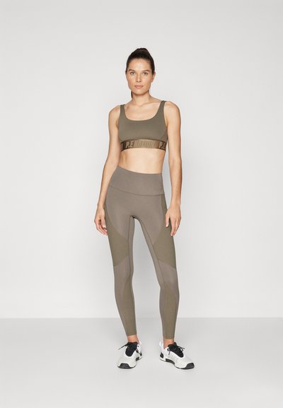 Olivengrønn sport-bh og høyhåndte leggings med teksturerte paneler og logo-bånd. Modell som har på seg svarte og hvite treningssko.