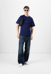 Camiseta de algodón de color azul marino con un pequeño detalle bordado, combinada con vaqueros de denim azul de corte holgado y zapatillas blancas; complementada con un bolso negro.