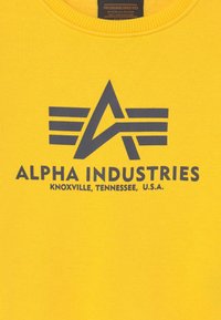 Felpa gialla con un logo grafico blu scuro che presenta ali stilizzate e il testo "ALPHA INDUSTRIES KNOXVILLE, TENNESSEE, U.S.A."