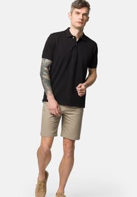 Czarna koszulka polo z klasycznym kołnierzykiem; krótkie rękawy; połączona z beżowymi szortami; gładki materiał; model ma tatuaż na lewym ramieniu.