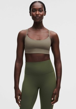 Frau trägt einen beigen Sport-BH und olivgrüne, hoch taillierte Leggings, steht vor einem schlichten, hellen Hintergrund.