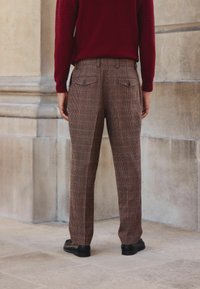 Brown plaid trousers with a tapered fit, featuring two back pockets and a smooth texture, paired with a burgundy sweater. Black shoes visible.