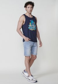 Mornarska majica bez rukava s živopisnom grafikom plaže, u kombinaciji sa svijetlim trapericama. Model nosi bijele i sive tenisice, stojeći s jednom rukom u džepu.