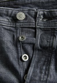Nærbillede af sorte denimjeans med tre metalknapper, hvoraf den ene er graveret med "aapreI", og en flossede knapphul i linningen.