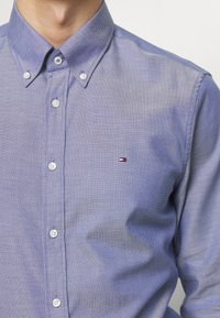 Camicia button-up di colore azzurro chiaro con un motivo texturizzato, dotata di colletto a punta, bottoni bianchi e un piccolo logo sul petto.