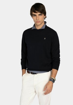 Joven con un suéter negro sobre una camisa gris con cuello, pantalones blancos y un reloj con pulseras en su muñeca izquierda, de pie con las manos en los bolsillos.