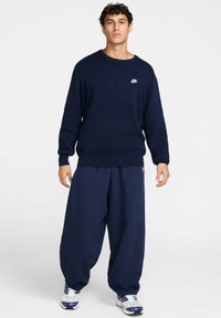 Ναυτικό μπλε πλεκτό πουλόβερ με στρογγυλή λαιμόκοψη και ριμπ μανσέτες, με μικρό λευκό λογότυπο Nike στο στήθος, σε συνδυασμό με φαρδιά παντελόνια. 