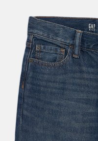 Σκούρο μπλε τζιν με κλασικό σχέδιο, πέντε τσέπες και πορτοκαλί ραφές. Η ετικέτα αναγράφει "GAP KIDS." Ομαλή υφή.