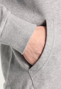 Mano parcialmente dentro del bolsillo delantero de una sudadera gris claro con textura de tela y costuras visibles.