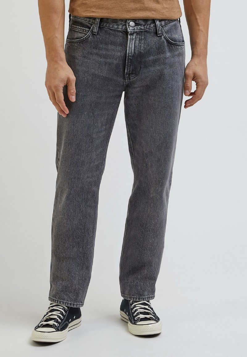 Homme portant un jean noir délavé à jambe droite et des baskets en toile noires avec des lacets blancs, debout devant un fond uni.