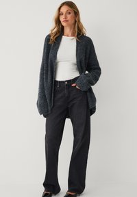 Frau in einer dunkelgrauen, oversized gestrickten Strickjacke, einem weißen, enganliegenden Langarmoberteil, lockeren schwarzen Jeans und schwarzen Schuhen vor einem schlichten Hintergrund.