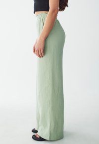 Pantalones de pierna ancha en verde claro, texturizados, con una cinturilla elástica. Combinados con sandalias negras, que destacan un diseño fluido y suave.