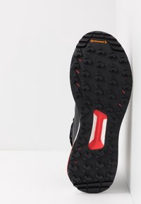 Semelle de chaussure de course noire en caoutchouc texturé pour une meilleure adhérence, avec un accent rouge et une semelle intermédiaire blanche. Conçue pour offrir une traction sur diverses surfaces.