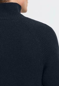 Maglione lavorato a maglia di un blu scuro, con un alto colletto, caratterizzato da una texture a coste verticali e maniche raglan. Materiale morbido, simile alla lana.
