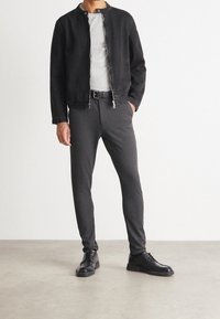 Man in een zwarte jas, lichtgrijs t-shirt, donkergrijze slim-fit broek, zwarte riem en zwarte leren schoenen die op een grijze vloer staat.