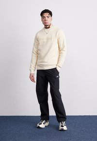 Sweatshirt galben deschis, cu logo-ul „Timberland” embosat, asortat cu pantaloni negri cu buzunare laterale și logo-ul New Balance. Adidași negri și albi.