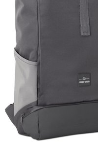 Rucsac gri cu un material texturat, partea de jos netedă gri în contrast, buzunar lateral și un eticheta de marcă neagră aproape de vârf.