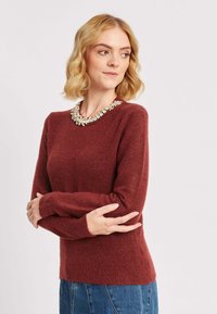 Burgundijas krāsas adīta džemperis ar garām piedurknēm un apaļu kakla izgriezumu, ar dekoratīvu dārgakmeņu apkakles detaļu. Mīksta tekstūra, brīva piegriezuma.