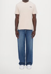 Camiseta de algodón beige con logo negro en el pecho, combinada con vaqueros azules de pierna ancha y zapatillas blancas. Corte sencillo y relajado. Sin patrones visibles.