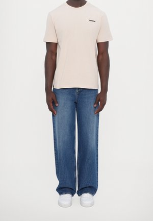 Beige katoenen T-shirt met zwart logo op de borst, gecombineerd met blauwe wijd uitlopende jeans en witte sneakers. Simpel, losse pasvorm. Geen zichtbare patronen.