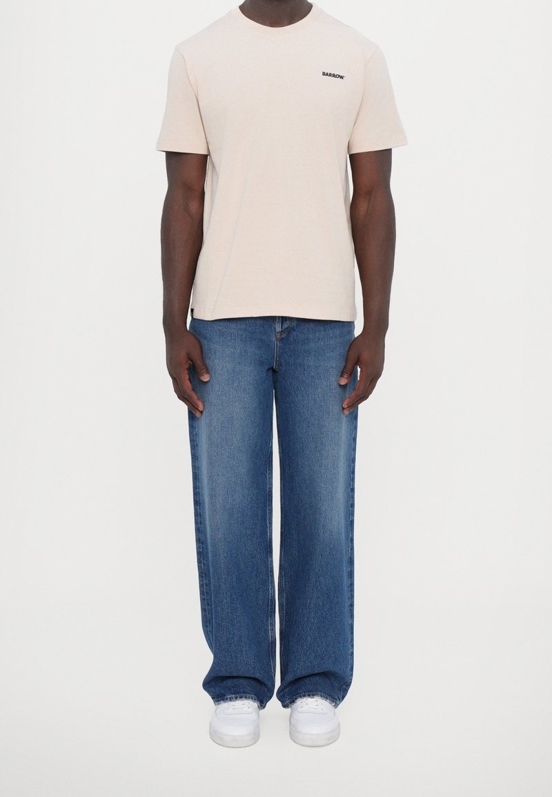 Camiseta de algodón beige con logo negro en el pecho, combinada con vaqueros azules de pierna ancha y zapatillas blancas. Corte sencillo y relajado. Sin patrones visibles.