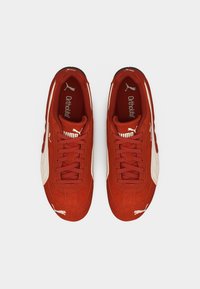 Ζευγάρι κόκκινα σουέντ παπούτσια Puma με λευκή ρίγα και λογότυπο, δεμένα με κορδόνια, φαίνονται από πάνω σε απλό φόντο.