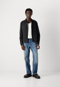 Svart knappskjorte med to frontlommer, hvit ribbestrikket tanktop, lyseblå denimjeans og svarte snøresko.